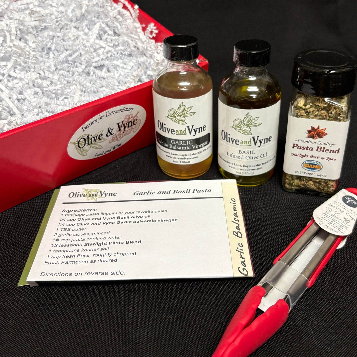 Garlic and Basil Pasta recipe card beside Garlic White Balsamic, Basil Infused Olive Oil, Pasta Blend seasoning, and mini tongs.