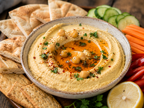 A bowl of garlic chili lemon hummus on a platter with pita, crackers and fresh vegetables.